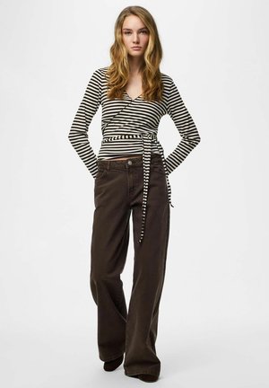 Giovane donna con capelli biondi lunghi che indossa una maglia avvolgente a righe bianche e nere e pantaloni ampi scuri, in piedi con le mani dietro la schiena.