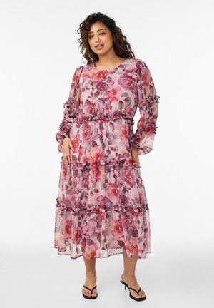 Žena v dlhorukávových, stredne dlhých ružovo-fialových kvetovaných šatách s volánikovými detailmi, stojaca s rukami pri bokoch na bielom pozadí.