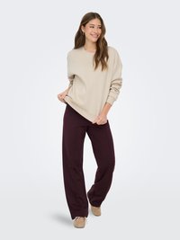 Femme souriante aux longs cheveux portant un sweat beige surdimensionné, un pantalon large violet foncé et des ballerines beiges, debout devant un fond uni.