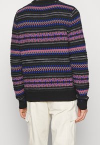 Henkilö, joka pitää päällä mustaa, purppuraista ja sinistä kuviollista neuletta sekä vaaleita cargo-housuja, seisoo selin yksinkertaista taustaa vasten.