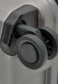 Rueda de plástico negro duro unida a un cuerpo de equipaje plateado texturizado, con un diseño redondeado y detalles visibles del mecanismo del eje.