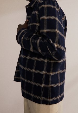 Person trägt eine marineblaue und weiße Karierten Jacke und richtet mit einer Hand den Kragen vor einem einfarbigen, hellen Hintergrund.