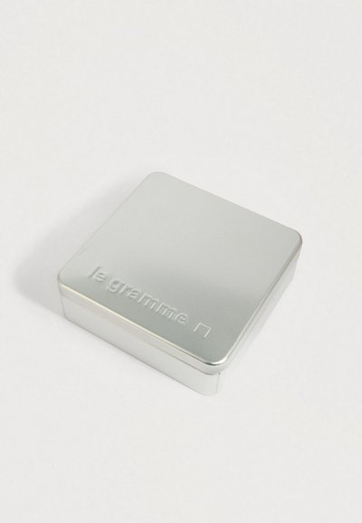 Kwadratowe srebrne metalowe pudełko z zaokrąglonymi krawędziami, wytłoczone z napisem "le gramme" i małym kwadratem na wieczku, umieszczone na białej powierzchni.