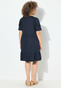 Laivansininen lyhythihainen mekko, jossa on frillahelma, istuva yläosa ja löysä hame. Tasainen kangas ja koristeelliset olkainyksityiskohdat.