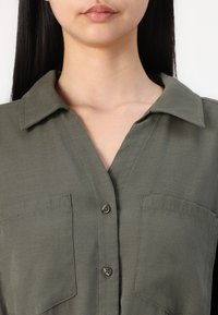 Camisa de botão verde oliva com colarinho, dois bolsos frontais e tecido texturizado. Apresenta um design clássico com um ajuste fluido.