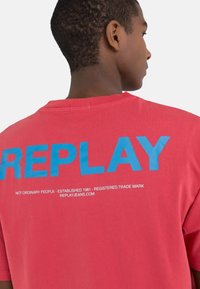 Punainen puuvillapaita, jossa on iso sininen "REPLAY" -teksti selässä ja lisävalkoinen teksti sen alla. Lyhyet hihat, rento istuvuus.