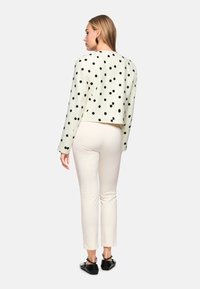 Gekürzter, cremefarbener Pullover mit Polka-Dots und langen Ärmeln, kombiniert mit enganliegenden, hellen Hosen und schwarzen Schuhen. Der Stoff wirkt strukturiert.