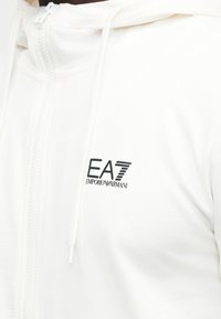 White zip-up hoodie with "EA7 Emporio Armani" logo in black on the chest and drawstrings hanging from the hood.
