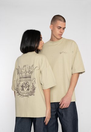 Beige oversized t-shirts van katoen met korte mouwen, met een achterafbeelding van een kroon en lettering, en een tekstdetail aan de voorkant.