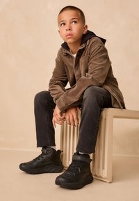 Čierne vysoké tenisky s gumovou podrážkou, šnúrkami a detailom s remienkom. Nosené s tmavými džínsami a hnedou kordurovou košeľou cez čiernu mikinu s kapucňou.