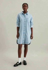 Giovane donna che indossa un vestito camicia di jeans azzurro chiaro, calze bianche e mocassini neri, in piedi contro uno sfondo chiaro e uniforme.