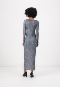 Long-sleeve, figure-hugging dress in green, featuring a floral pattern with purple accents. Smooth texture and full-length design, paired with black heels.