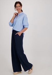 Lichtblauwe gestreepte overhemd met knopen, marineblauwe geplooide wijde broek en lichtbeige espadrilles. De look bevat grote creool oorringen.