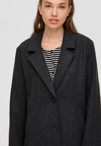 Dunker grauer Wollmantel mit einem Reverskragen, zwei schwarzen Knöpfen und seitlichen Taschen. Über einem schwarz-weiß gestreiften Oberteil getragen.