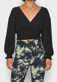 Top nero cropped con maniche lunghe e larghe e scollatura a V profonda; vita arricciata con bordi frangiati. Abbinato a pantaloni tie-dye verdi e neri.