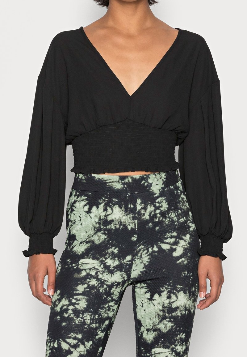 Top nero cropped con maniche lunghe e larghe e scollatura a V profonda; vita arricciata con bordi frangiati. Abbinato a pantaloni tie-dye verdi e neri.