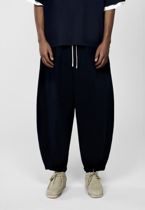 Personne debout portant un pantalon noir ample avec des cordons blancs, une chemise foncée sur une manche blanche, et des chaussures en daim beige, sur un fond uni.