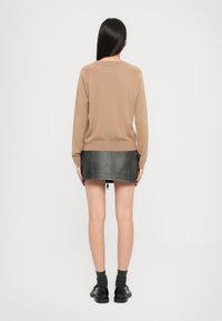 Kvinne med langt svart hår iført en beige genser, svart miniskjørt, grå sokker og svarte sko, stående med ryggen til mot hvit bakgrunn.