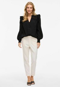 Blouse noire à manches bouffantes avec détail de volants aux épaules, devant à boutons, associée à un pantalon crème fuselé et des sandales noires à bout ouvert.