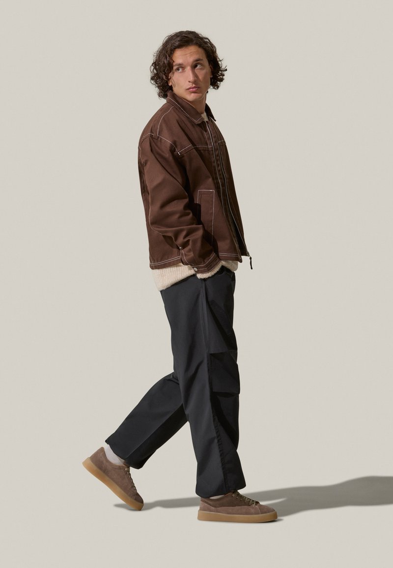 Giacca di denim marrone con cuciture bianche a contrasto, indossata sopra un maglione beige; abbinata a pantaloni cargo neri e sneakers marroni.