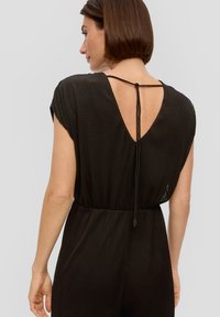 Zwarte geribbelde jumpsuit met een diepe V-rug, korte mouwen en een getailleerde taille. Inclusief een striksluiting aan de achterkant en subtiele textuur in de stof.
