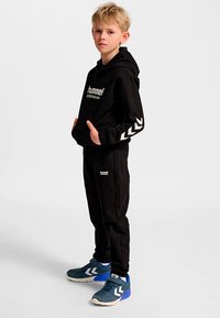 Sort hoodie og joggingbukser sæt med logo og hvide chevron-accenter. Drengen har blå træningssko med hvide detaljer.