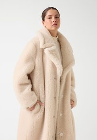 Donna che indossa un lungo cappotto di shearling soffice color crema, con le mani nelle tasche su uno sfondo bianco neutro.