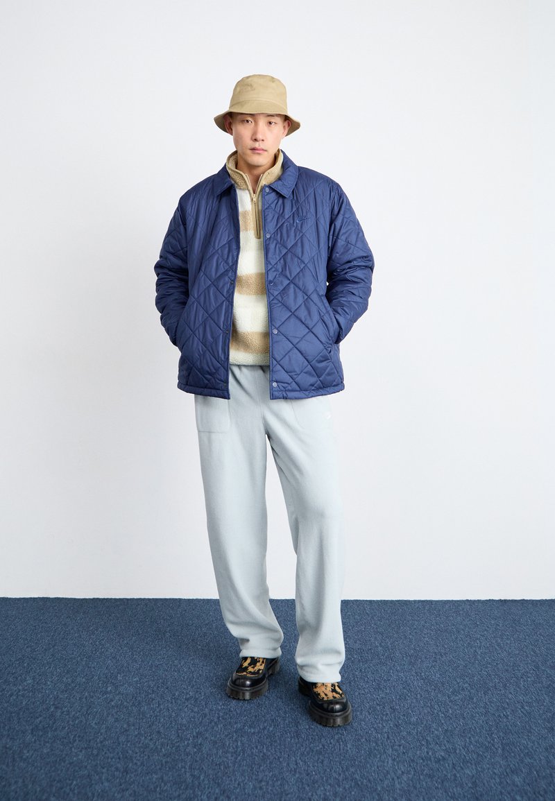 Veste matelassée bleu marine sur un polaire beige rayé, associée à un pantalon bleu clair et des chaussures à plateforme noires avec un imprimé léopard. Chapeau seau beige.