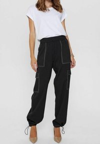 Pantalon cargo noir avec une taille élastique, deux poches avant et des détails de couture blanche. Bords resserrés avec cordons de serrage. Porté avec un t-shirt blanc.