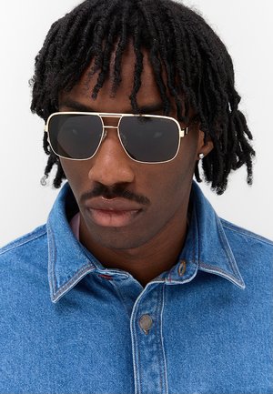 Uomo con capelli attorcigliati che indossa occhiali da sole rettangolari scuri, un piccolo orecchino e una giacca di jeans blu con il colletto alzato.