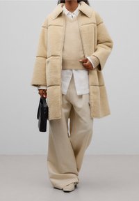 Beige shearlingjacka med vida ärmar, matchande beige stickad tröja, vit skjorta under, krämfärgade byxor och en svart rektangulär väska.