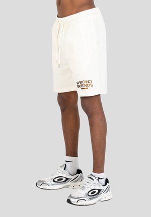 Uomo che indossa pantaloncini bianchi con coulisse con la scritta "WRONG FRIENDS", calzini bianchi Nike e scarpe da ginnastica bianche e nere, in piedi su sfondo grigio.