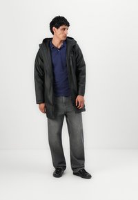 Giacca nera impermeabile con cappuccio, abbinata a una camicia blu a pois e jeans grigi, in piedi su uno sfondo bianco.