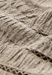 Tessuto strutturato beige con strati arricciati e motivo intrecciato lungo il bordo. Materiale morbido e leggero con una finitura naturale.