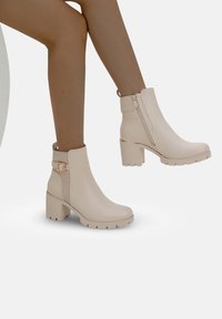 Botas de tobillo beige con un acabado suave, cremallera lateral, una correa decorativa con hebilla y un tacón grueso con surcos para mayor agarre.
