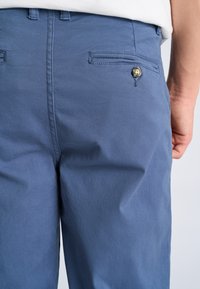 Pantaloni di cotone azzurro chiaro con texture liscia, tasca posteriore con dettaglio bottone e passanti per cintura. Design semplice con taglio classico.