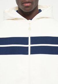 Homme portant un sweat à capuche blanc zippé avec deux bandes horizontales bleu marine sur la poitrine et une capuche.