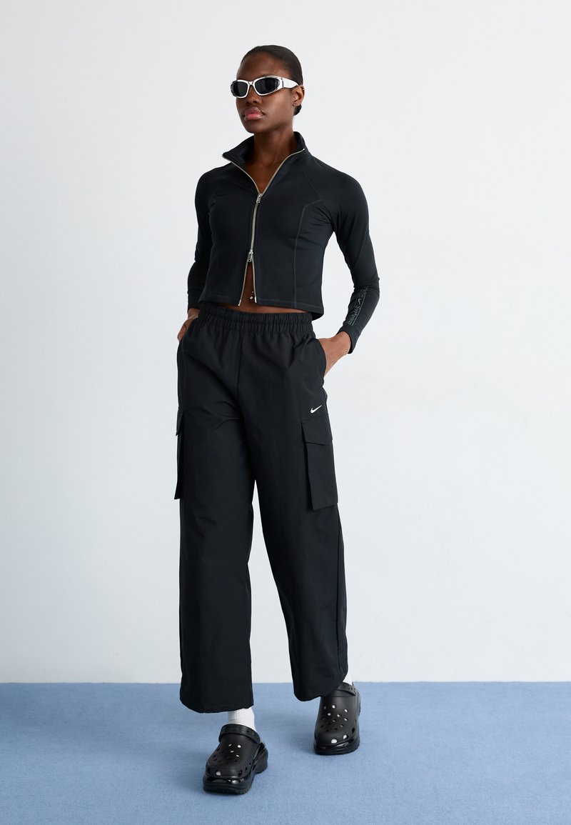 Schwarze, kurze Reißverschlussjacke mit langen Ärmeln, kombiniert mit weiten schwarzen Cargo-Hosen mit Taschen. Schwarze Clogs runden das Outfit ab.