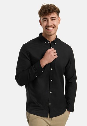 Nuori mies hymyilee, yllään musta napillinen paita ja beiget housut, säätää kaulusta yhdellä kädellä, yksinkertaisen vaalean taustan edessä.