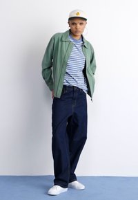 Veste bombardier verte sur un t-shirt rayé bleu et blanc, associée à un jean en denim sombre et des baskets blanches, surmontée d'une casquette claire.
