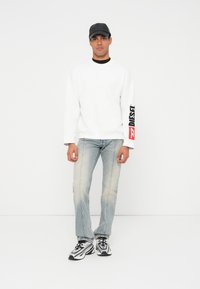 Felpa bianca a maniche lunghe con scollo nero e logo "DIESEL" rosso sulla manica, abbinata a jeans chiari strappati e sneakers bianche e nere.