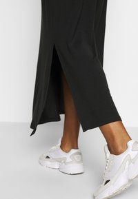 Robe maxi noire en jersey avec une fente latérale, assortie à des baskets blanches de sport avec un design texturé et des accents de logo.