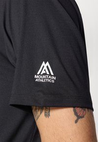 Juodas sportinis marškinėliai trumpomisRankovėmis, su balta "Mountain Athletics" logo kairėje rankovėje ir lygiu, tekstūruotu audiniu.