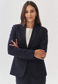 Námořnický modrý blazer s přiléhavým střihem, zapínáním na jeden knoflík a výstřihy ve tvaru klopy, nošený přes světlý top. Hladká textura tkaniny.