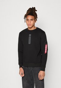 Sudadera negra con cuello redondo, puños y dobladillo acanalados. Presenta un logo central y un bolsillo lateral con cremallera con un detalle de etiqueta roja.
