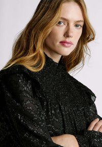 Blusa nera con texture, colletto alto arricciato, spalle a volant e intricate decorazioni di perline su tutto il tessuto.