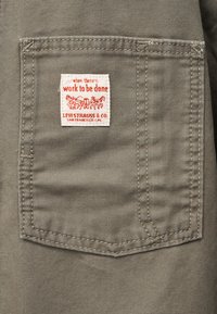 Τσέπη από καφέ χακί ύφασμα με λευκή και κόκκινη ετικέτα Levi Strauss & Co. που γράφει «όταν υπάρχει εργασία να γίνει» και με γραφικό που απεικονίζει άμαξα με άλογα.