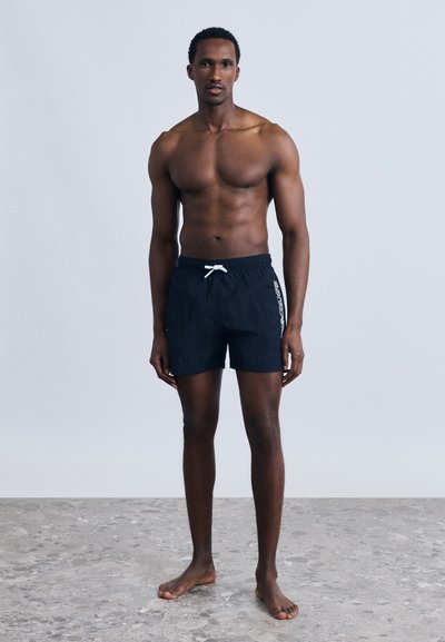 Άνδρας χωρίς πουκάμισο, όρθιος ξυπόλητος σε γκρι δάπεδο, φοράει μαύρο μαγιό με λευκό κορδόνι και λογότυπο στο πλάι, μπροστά σε απλό φωτεινό φόντο.