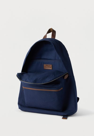 Σακίδιο πλάτης από καμβά σε ναυτικό μπλε χρώμα με καφέ δερμάτινη λαβή, μπροστινή τσέπη με φερμουάρ και κύριο διαμέρισμα μερικώς ανοιχτό που αποκαλύπτει το εσωτερικό λογότυπο.