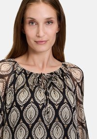 Schwarze und cremefarbene blouse mit gemustertem Design, lockerer Passform, runden Ausschnitt und Bindedetail. Transparenter Stoff mit paisley-Motiven durchgehend.